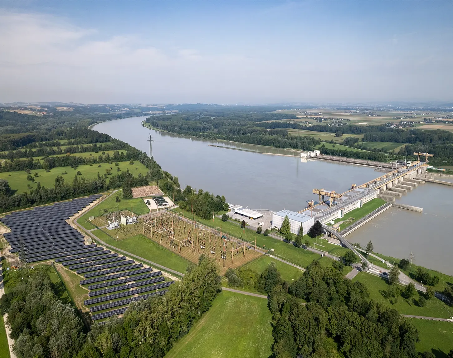 Das Kraftwerk Wallsee-Mitterkrichen von oben. Inklusive Batteriespeicher und Photovoltaikanlage. Die Umgeben ist grün und die Sonne scheint.