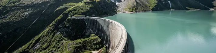 Ein Blick auf den Staussee bei Kaprun. Im Vordergrund sind Wiesen und Blumen zu sehen. Im Hintergrund schneebedeckte Berge.